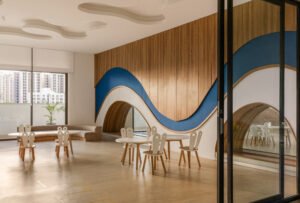 Classroom interior at Vedanya School featuring blue wave wood wall cladding, low arched peekaboo cutout window into adjacent space, round study tables, and organic cloud ceiling panels.