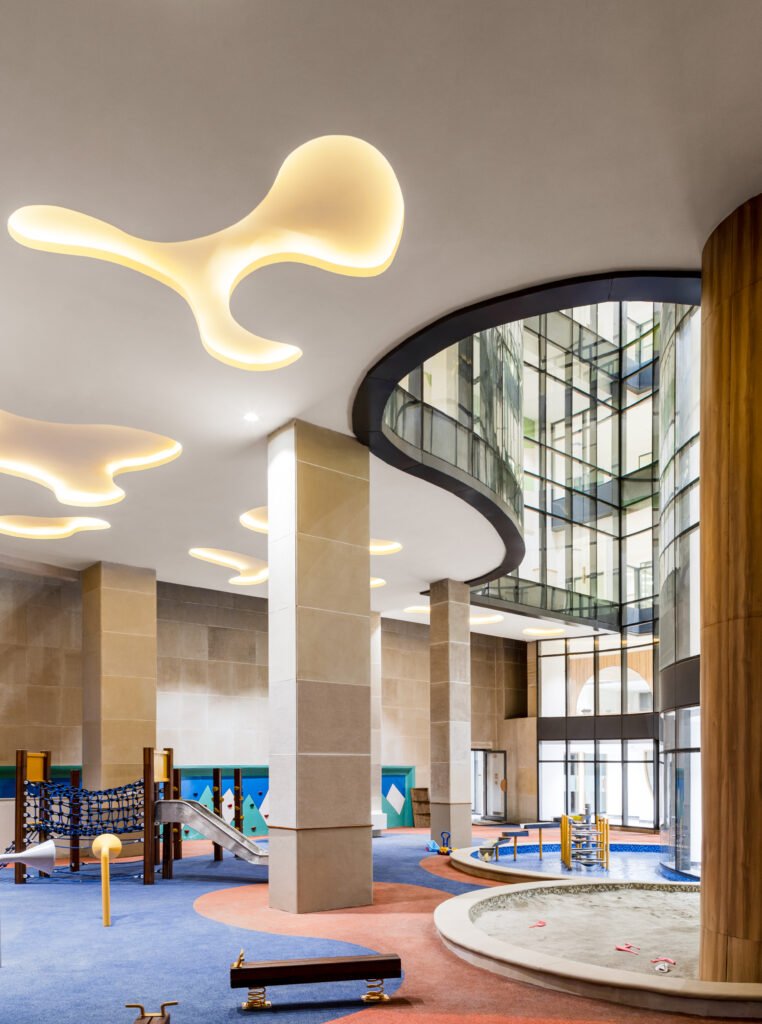 Covered outdoor play space under double-height volume at Vedanya School, featuring play climbing nets, slide, sand pit, water play station, and organic cloud ceiling light coves.