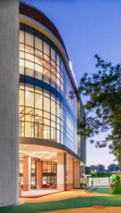 Evening exterior photography of Vedanya School showcasing multi-story curved glass curtain wall, vertical circulation volume, stilted play area, and colorful outdoor play turf.