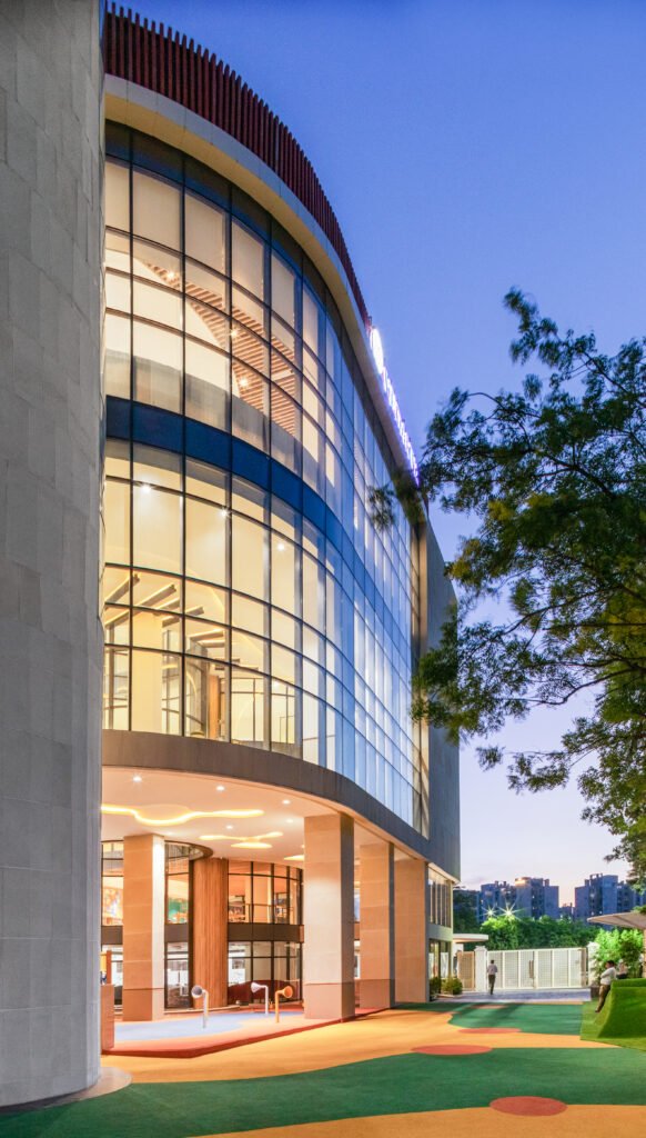 Evening exterior photography of Vedanya School showcasing multi-story curved glass curtain wall, vertical circulation volume, stilted play area, and colorful outdoor play turf.