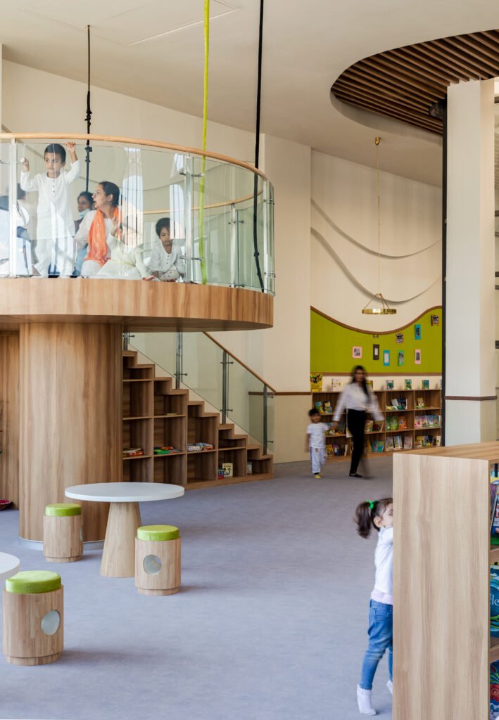 Custom wooden staircase with built-in shelf storage leading to a glass-balustraded mezzanine reading loft in the Vedanya School library.