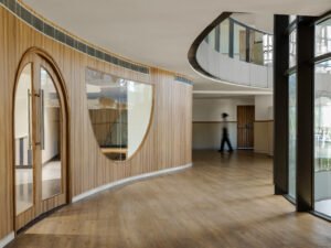 Modern school hallway featuring curved wood-veneered walls, arched glass door entrance, custom teardrop-shaped window cutout, and polished light wood floors at Vedanya School.