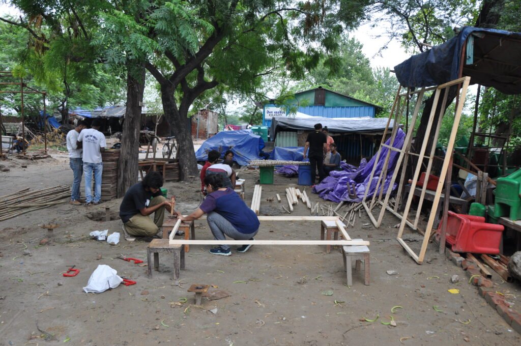 An outdoor carpentry workspace where participants assemble rectangular wooden frames supported on small wooden stools amidst scattered timber and tools.