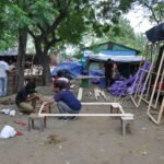 An outdoor carpentry workspace where participants assemble rectangular wooden frames supported on small wooden stools amidst scattered timber and tools.