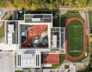 Bird's-eye aerial view showing the square two-storey building ring, central play yard, extensive green roofs, rooftop solar panels, and outdoor sports fields.